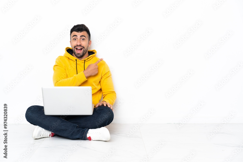 Caucasian man sitting on the floor with his laptop surprised and pointing side Caucasian man sitting on the floor with his laptop surprised and pointing side