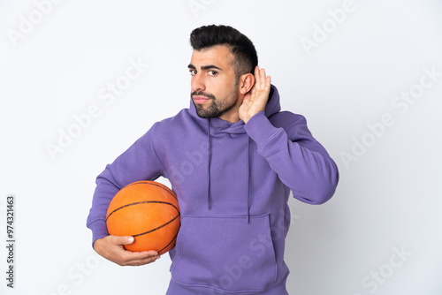 Man playing basketball over isolated white wall listening to something by putting hand on the ear