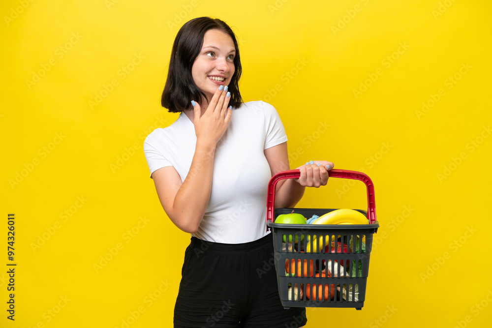 Young woman holding a shopping basket full of food isolated on yellow background looking up while smiling Young woman holding a shopping basket full of food isolated on yellow background looking up while smiling