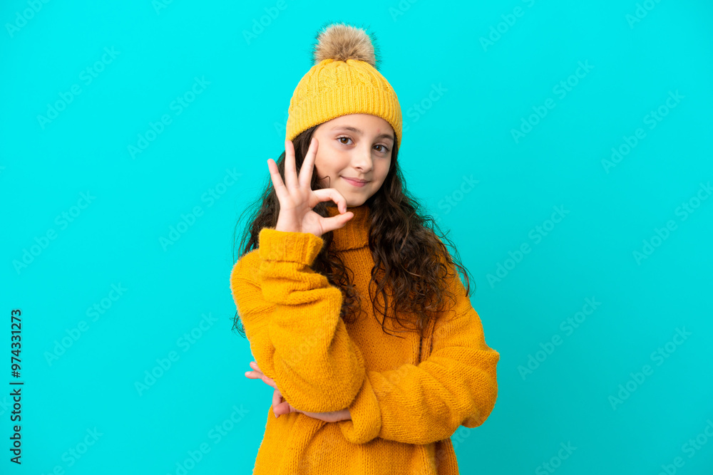 Little caucasian girl wearing winter hat isolated on blue background showing ok sign with fingers Little caucasian girl wearing winter hat isolated on blue background showing ok sign with fingers
