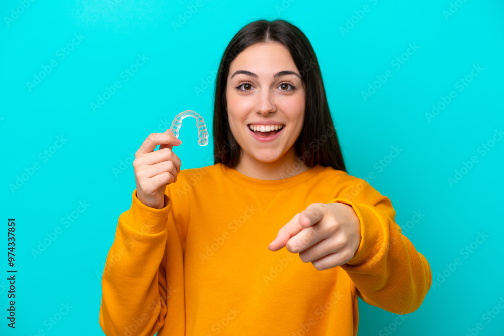 Young caucasian woman holding invisible braces isolated on blue background surprised and pointing front Young caucasian woman holding invisible braces isolated on blue background surprised and pointing front