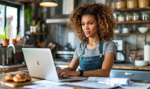 A young woman with curly hair working on a laptop in a cozy kitchen. She is wearing a gray t-shirt and denim overalls, focused on her task.