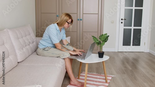 Side shot of a young tattooed woman in eyeglasses and a polo using a silverish laptop standing on a three legged table while sitting on a light couch in a living room.