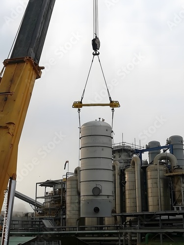 A large white tank with a conical base and flanges is lifted by a yellow crane with a long boom against a blue sky. Industrial buildings are visible in the background. photo