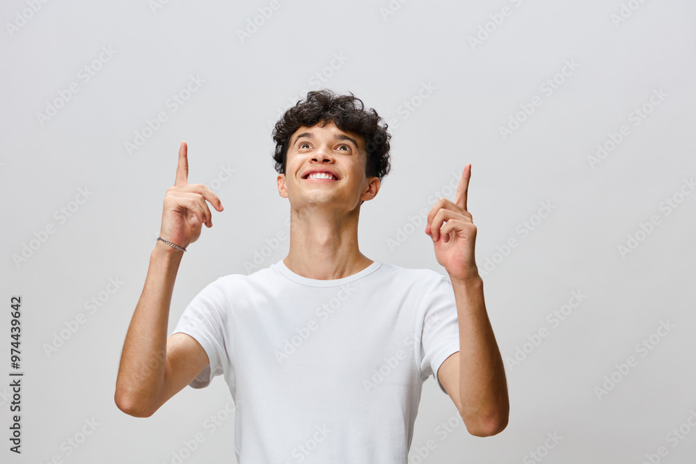 Young man smiling while pointing upwards against a light gray background, expressing excitement and energy A joyful moment highlighting positivity and enthusiasm Young man smiling while pointing upwards against a light gray background, expressing excitement and energy A joyful moment highlighting positivity and enthusiasm