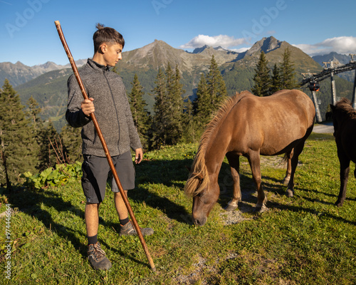 A young person with a stick interacts with a horse on a mountain hillside with scenic views during daylight