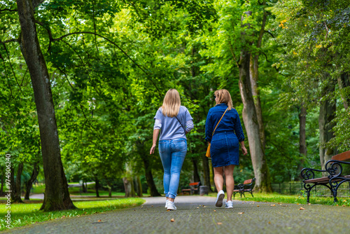 Two young beautiful smiling women dressed casual walking in city park on sunny day, trees in background. Meeting of friends. Walk in park. Back view