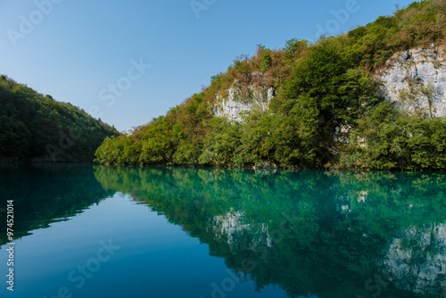 Emerald-hued reflections dance on the tranquil surface of Plitvice Lakes, framed by lush greenery and steep rocky cliffs, inviting visitors to immerse themselves in nature's beauty.