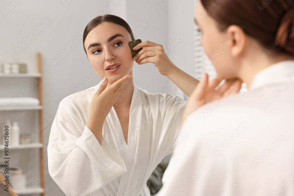 Beautiful young woman doing facial massage with gua sha tool near mirror at home Beautiful young woman doing facial massage with gua sha tool near mirror at home