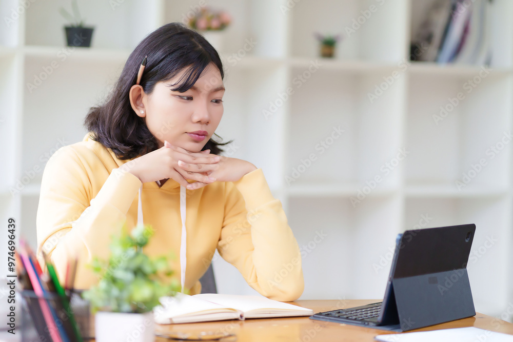 A young woman in a yellow hoodie is sitting at a table, typing on her laptop. She is working in a bright room with large windows, surrounded by natural light and greenery outside