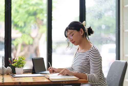 Free Download A young woman wearing glasses and a striped shirt is sitting at a desk, writing in a notebook. She appears focused, with an organized indoor setting around her and shelves in the background - GFXLoad