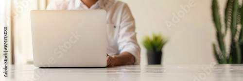 Business professional in a white shirt works on a laptop in a well-lit office with natural sunlight and minimalistic decor, including a small plant in the background.