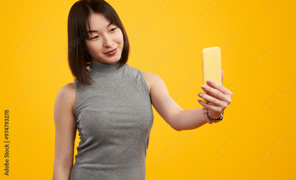Young woman holding a smartphone and taking a selfie with a cheerful expression on a vibrant yellow background. Young woman holding a smartphone and taking a selfie with a cheerful expression on a vibrant yellow background.
