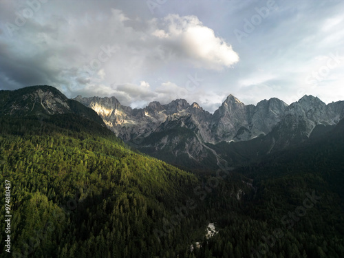 mountain village nestled at the base of towering, rugged mountains covered in dense forests. Sunlight breaks through the clouds, casting beautiful shadows over the vibrant green fields and small, scat