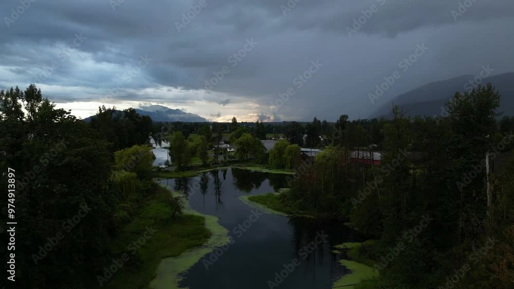 Aerial view of a rural landscape with a pond, trees, fields and mountains in the background, under a dramatic, stormy sky. British Columbia, Canada. Aerial view of a rural landscape with a pond, trees, fields and mountains in the background, under a dramatic, stormy sky. British Columbia, Canada.