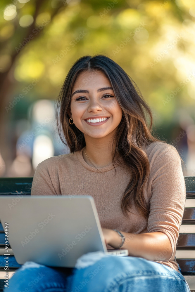 A joyful young Hispanic female college student e-learning on her laptop at campus, sitting on a bench and smiling warmly at the camera.
