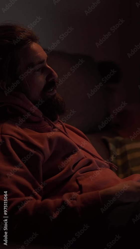 Vertical. Caucasian man with beard sitting quiet on sofa changing channel with TV remote control. Young casual millennial male smile enjoying movie, documentary or series relaxing comfort at night Vertical. Caucasian man with beard sitting quiet on sofa changing channel with TV remote control. Young casual millennial male smile enjoying movie, documentary or series relaxing comfort at night