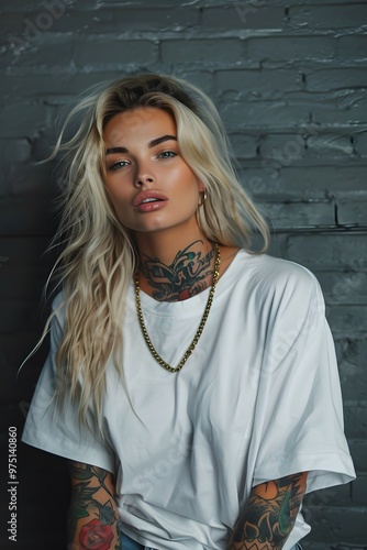 A beautiful girl with tattoos stands in front of an oversize white t-shirt mockup with a light concrete backdrop