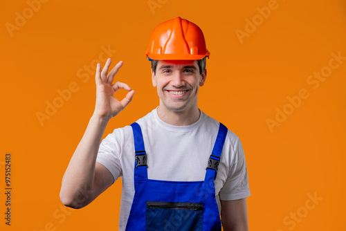 Positive builder in helmet, protective uniform makes OK okay hand sign on orange