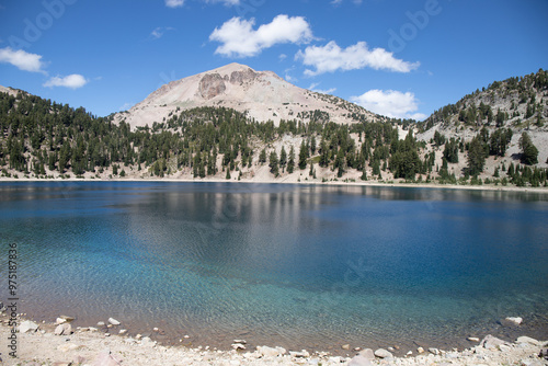 A serene lake reflects the surrounding pine tree-covered mountains under a bright blue sky with scattered clouds. Emerald Lake is located in Lassen Volcanic National Park