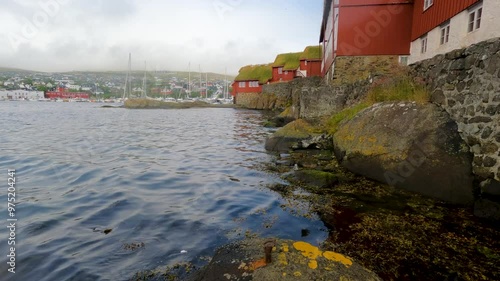 Red Scandinavian style buildings with grass rooftops on the rocky shoreline of the North Atlantic Ocean. Large ships are moored in the harbor of Tórshavn, the Capital city of the Faroe Islands.  photo