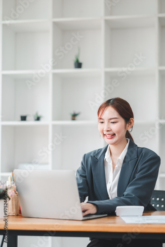 A young professional woman in a blazer working on a laptop in a modern office with white shelves in the background.