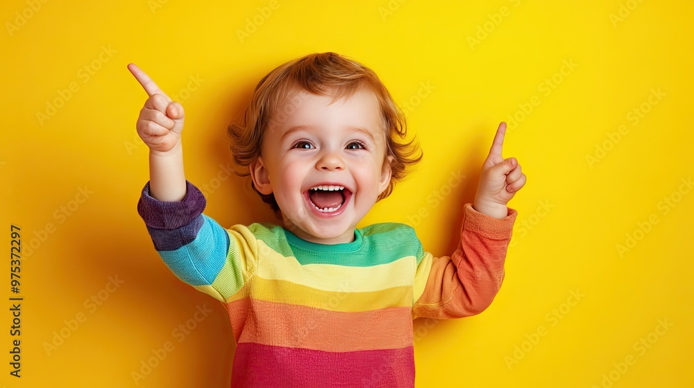 A cheerful toddler in a rainbow sweater points upwards with both hands against a bright yellow background. A cheerful toddler in a rainbow sweater points upwards with both hands against a bright yellow background.