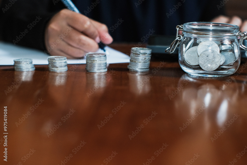 Saving concept with pile of the coins and coins on the jar glasses and a hand holding pen taking note the amount of money with calculator in the wooden table, Savings and investing concept. Saving concept with pile of the coins and coins on the jar glasses and a hand holding pen taking note the amount of money with calculator in the wooden table, Savings and investing concept.