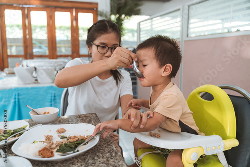 Mother feeding her child in a high chair with food on the table in a casual home setting.