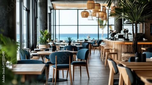 Modern restaurant interior with wooden tables and chairs, blue upholstery, and large windows overlooking a beach and ocean.