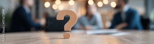 A wooden question mark stands prominently in focus while three blurred people are having a discussion in the background within an office setting.
