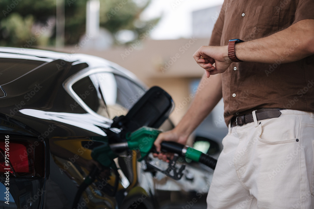 Fototapeta premium Low angle of male driver in casual clothes filling fuel into modern car with gas pump nozzle at station. Middle selection. Man looking at his watch