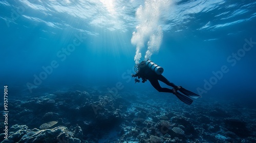 A scuba diver swims through the clear blue water over a coral reef with sunlight shining through the surface.