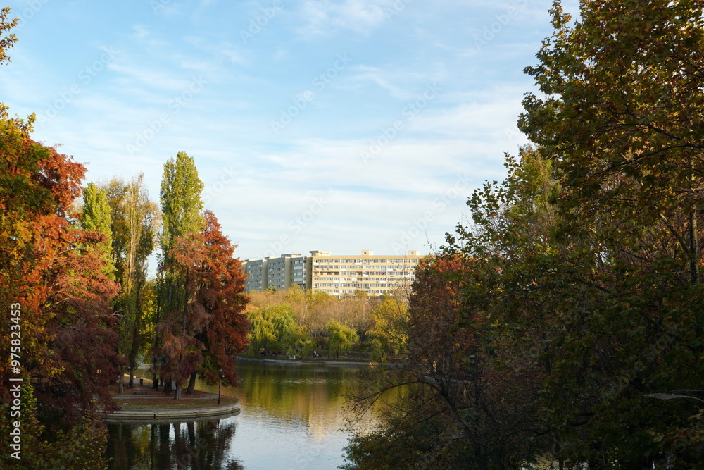 Fototapeta premium Beautiful autumn landscape with trees, lake and block of apartments in IOR park in Titan neighborhood in Bucharest, Romania