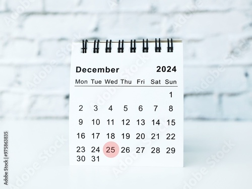 A minimalist and modern desktop calendar showing December 2025, with Christmas Day 25th highlighted by a red circle. It sits on a white table against a white brick wall