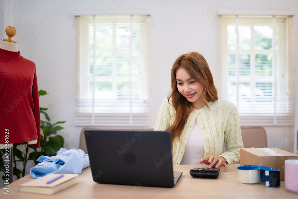 A young woman running an online clothing business is smiling while using her laptop at a desk. She is surrounded by clothing items and managing her orders. A young woman running an online clothing business is smiling while using her laptop at a desk. She is surrounded by clothing items and managing her orders.