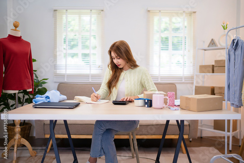 A young woman is sitting at a desk, writing in a notebook while working on her online clothing business. She appears focused, with laptops, clothing, and a mannequin nearby in a well-lit room