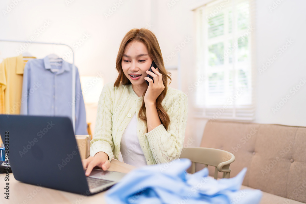 A young woman is talking on the phone while working on her laptop, managing her online clothing business. She is in a bright room A young woman is talking on the phone while working on her laptop, managing her online clothing business. She is in a bright room