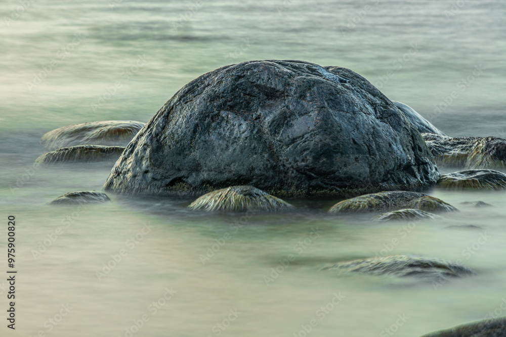 Obraz premium A serene close-up of moss-covered rocks gently submerged in calm coastal waters. The soft light and smooth water create a peaceful, almost ethereal atmosphere