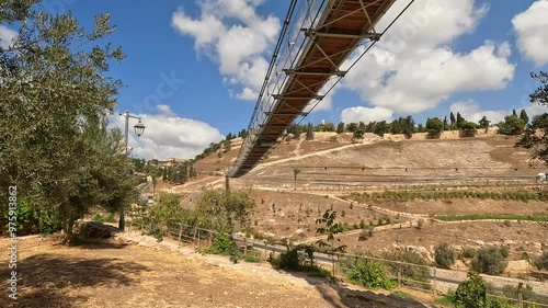 the suspension bridge over Gei Ben Hinnom near the Old City of Jerusalem, with the Mount of Olives and cloudy skies in the background. 