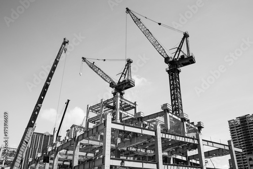 Black and white photo, Low-angle view of cranes and steel structures of building construction with a blue sky background.