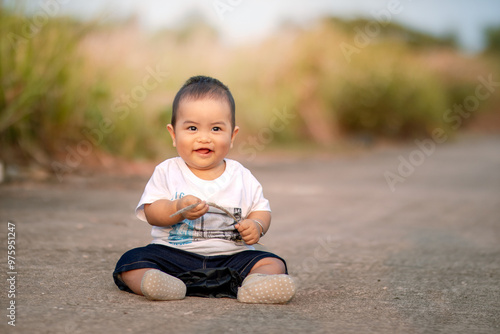 cute asian baby sitting on the road. baby boy holding wild grass while smiling sweetly wearing a white shirt towards the camera. photo atmosphere in the afternoon.