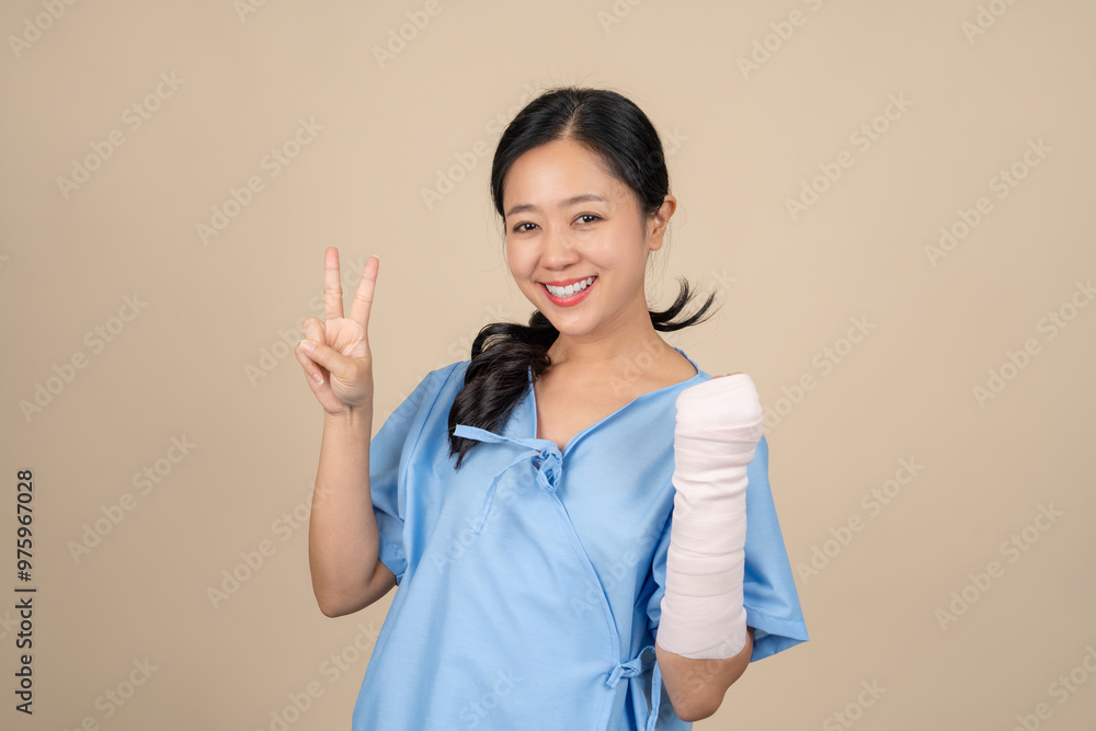 Beautiful Asian woman wearing patient gown and cast due to accident injury, her hand looks okay on cream background. Insurance concept. Beautiful Asian woman wearing patient gown and cast due to accident injury, her hand looks okay on cream background. Insurance concept.