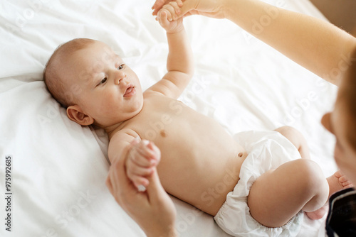 A mother interacts lovingly with her baby, holding their hands as they lie on a light-colored bed, creating a warm and intimate atmosphere in their home.