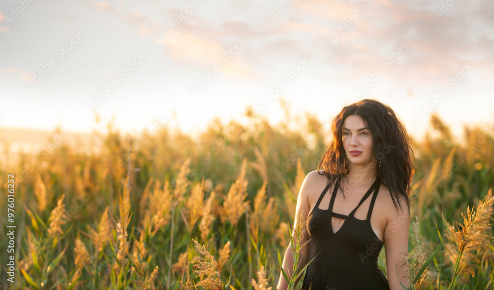 A girl with dark curly hair in a short dress among the grass on the shore of a pond at sunset. A girl with dark curly hair in a short dress among the grass on the shore of a pond at sunset.