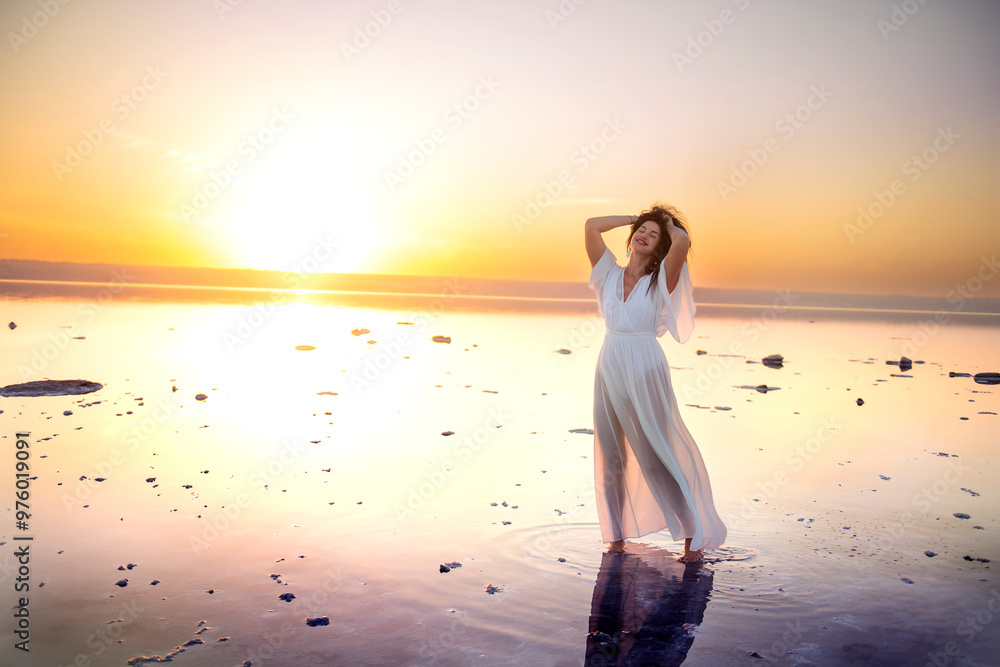 Fototapeta premium Beautiful girl with long dark curly hair in a long airy white dress stands in the water of a lake at sunset. mirror reflection of the sky in the surface of the lake. Fantastically beautiful view of a