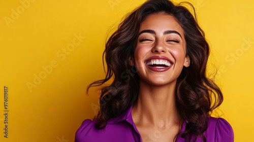 A beautiful woman with long brown hair is laughing while wearing a purple shirt and standing in front of a yellow background.