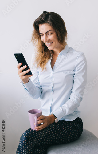 Smiling young female employee with cup sitting and using smartph photo