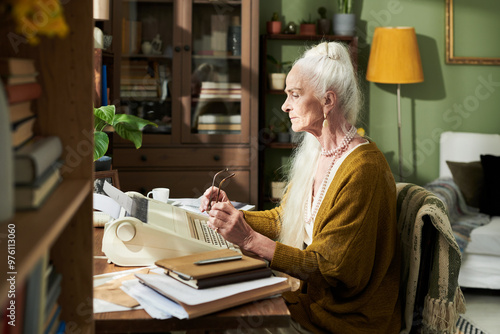 Elderly woman working on typewriter in cozy home office with bookshelves and green walls surrounding her. She holds a pair of glasses while concentrating on her work photo