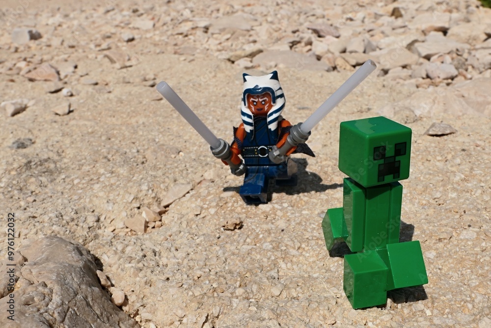 Naklejka premium ĹEGO Star Wars figure of female Jedi of Togruta race Ahsoka Tano hunting LEGO Minecraft explosive green mob with two lightsabers on sunlit summer rocky beach.